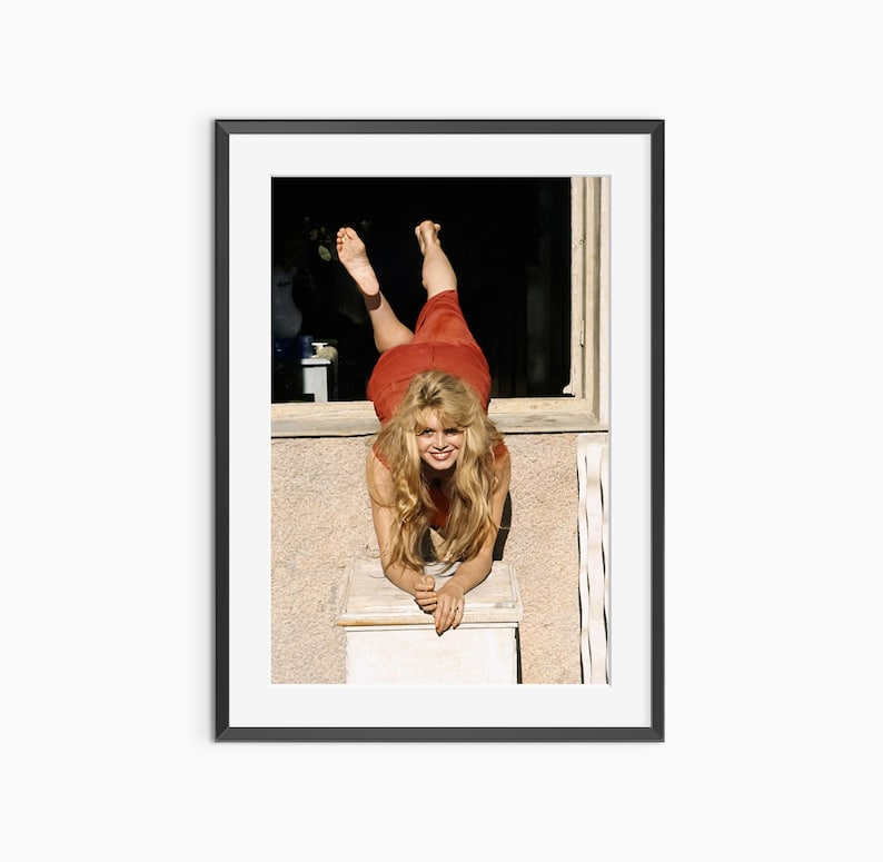 A woman in a red dress is leaning against a window sill, smiling and looking out at the camera.