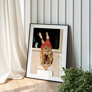 A framed photograph of a smiling woman in a red dress is displayed on a wooden floor next to a potted plant.