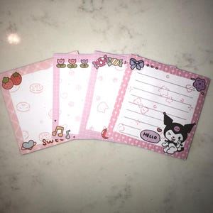 Op de afbeelding: Een stapel van vijf roze vierkante notitieblokken met verschillende ontwerpen. Elk notitieblok heeft een roze rand en een witte binnenkant met illustraties zoals aardbeien, donuts, muzieknoten en een stripfiguur met het woord "HELLO".