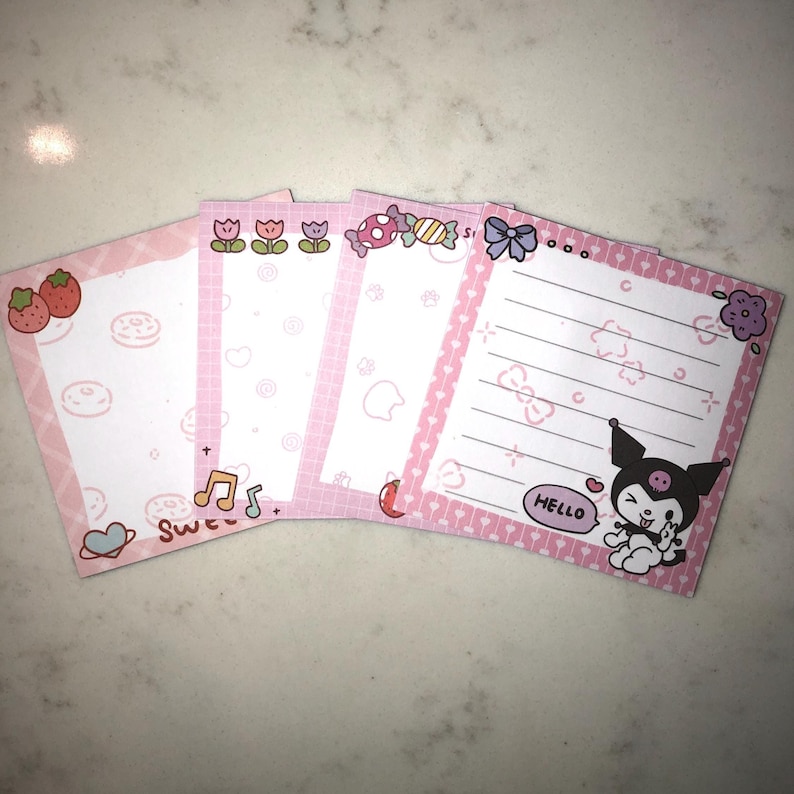 Op de afbeelding: Een stapel van vijf roze vierkante notitieblokken met verschillende ontwerpen. Elk notitieblok heeft een roze rand en een witte binnenkant met illustraties zoals aardbeien, donuts, muzieknoten en een stripfiguur met het woord "HELLO".