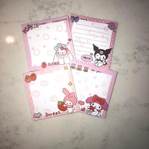 Op de afbeelding: Vier roze memoblokken met stripfiguren en ontwerpen. E&eacute;n blok heeft Hello Kitty met het woord "CAKE". Een ander heeft My Melody met een aardbei met het woord "Sweet". De derde heeft Kuromi met "HELLO". De vierde heeft My Melody met een hart.