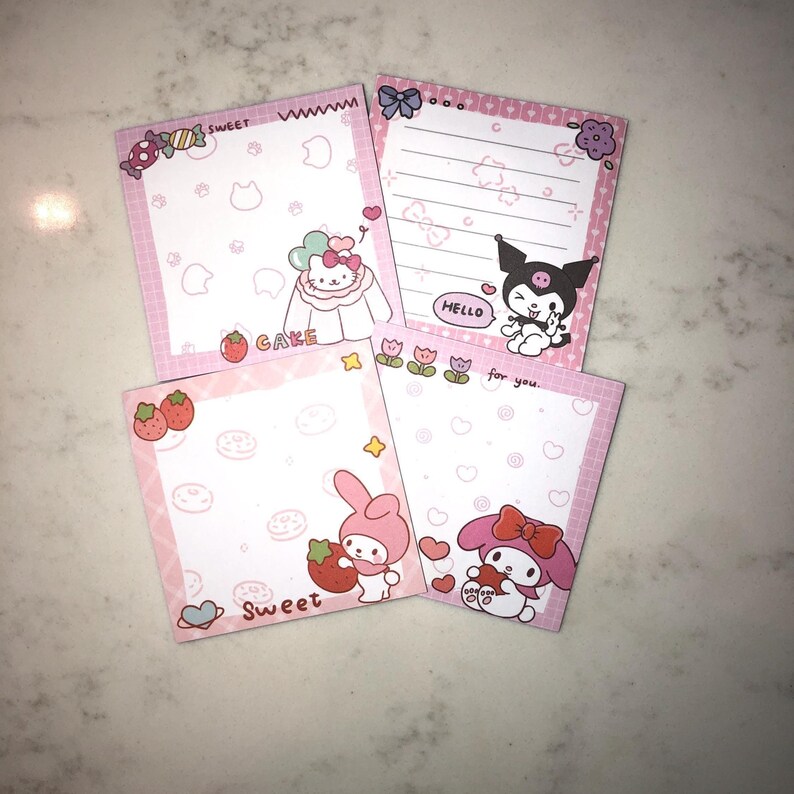 Op de afbeelding: Vier roze memoblokken met stripfiguren en ontwerpen. E&eacute;n blok heeft Hello Kitty met het woord "CAKE". Een ander heeft My Melody met een aardbei met het woord "Sweet". De derde heeft Kuromi met "HELLO". De vierde heeft My Melody met een hart.