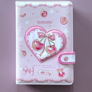 Puede incluir: Cuaderno rosa con diseño de cerezas en forma de corazón. La portada presenta un corazón con cerezas, lazos y las palabras "Fruit series" y "Cherry". El cuaderno tiene un cierre magnético.