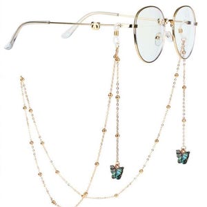 Könnte beinhalten: Goldfarbene Brille mit runden Gläsern und einer filigranen Kette. Die Kette hat kleine goldene Perlen und zwei Schmetterlingsanhänger mit schillernden türkis-schwarzen Flügeln. Die Brille hat einen dünnen Goldrahmen.