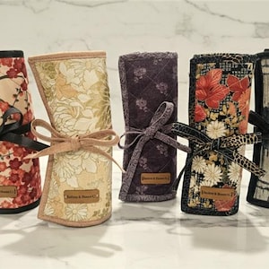 May include: Five fabric roll-up cases with various floral and patterned designs. Each case is tied with a ribbon bow. The cases feature a variety of colors, including red, cream, purple, and black.