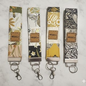 May include: Four fabric wrist keychains with floral and insect patterns. Each keychain has a different design, including bees, flowers, and leaves. Each keychain has a metal clasp and a leather tag that says "BBCo".