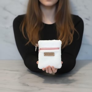 May include: A white, fluffy pouch with a pink zipper and a small metal tag. The pouch is held in front of a person. The tag reads "Boston & River Co."