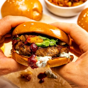 May include: A freshly made burger held in hands, featuring a sesame seed bun, a thick patty, and various toppings including red sauce, cucumber, and cilantro. Additional buns and a side dish are visible in the background.