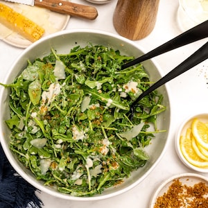 Op de afbeelding: Rucola salade in een grote witte kom, geserveerd met geraspte Parmezaanse kaas, croutons en dressing. Extra ingrediënten zijn citroen schijfjes, een houten pepermolen en een mes met een stuk kaas.