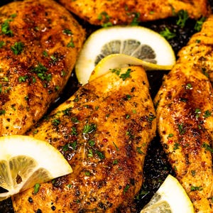 以下が含まれることがあります： ハーブとスパイスで味付けされ、レモンスライスで飾られた、調理された鶏むね肉のクローズアップ。鶏肉は黄金色で、暗い表面に盛り付けられています。美味しくて風味豊かな食事。