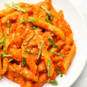 May include: A white plate of penne pasta in a rich orange sauce, topped with fresh green basil. The pasta is covered in a creamy sauce and sprinkled with grated cheese. A visually appealing and tasty dish.