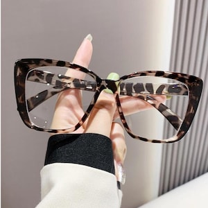 May include: Eyeglasses with a leopard print design. The frames are brown and black, with a square shape. The lenses are clear. The glasses are held up by a hand, showing the frame's pattern.