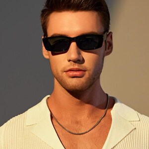 May include: A person wearing black rectangular sunglasses and a silver chain necklace. The person is wearing a white collared shirt with a textured pattern. The background is a neutral color.