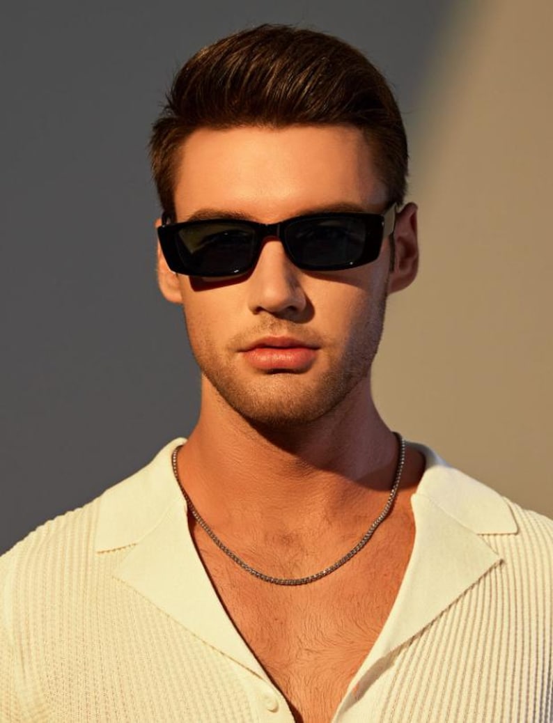 May include: A person wearing black rectangular sunglasses and a silver chain necklace. The person is wearing a white collared shirt with a textured pattern. The background is a neutral color.