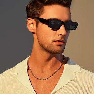 May include: A person wearing black rectangular sunglasses and a silver chain necklace. The person is wearing a white collared shirt. The background is a gradient of light and dark neutral tones.
