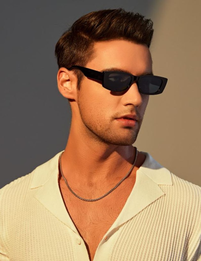 May include: A person wearing black rectangular sunglasses and a silver chain necklace. The person is wearing a white collared shirt. The background is a gradient of light and dark neutral tones.