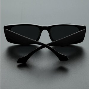 May include: Black sunglasses with a rectangular frame and dark lenses. The temples are thin and curve away from the frame. The sunglasses are set against a dark background.