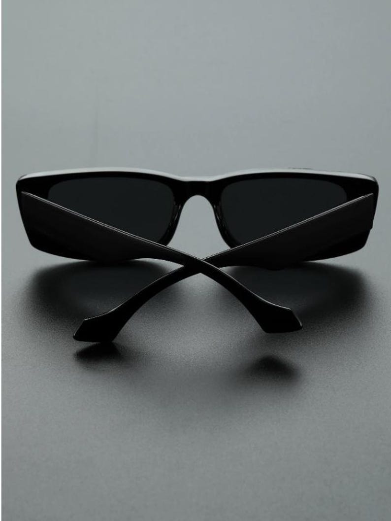 May include: Black sunglasses with a rectangular frame and dark lenses. The temples are thin and curve away from the frame. The sunglasses are set against a dark background.