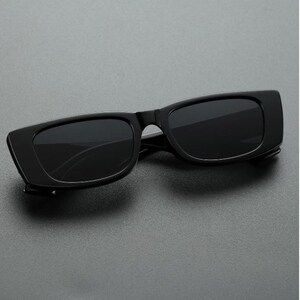May include: Black rectangular sunglasses with dark lenses. The sunglasses have a thick, solid black frame and a sleek, modern design. The lenses are dark, providing shade and protection from the sun.