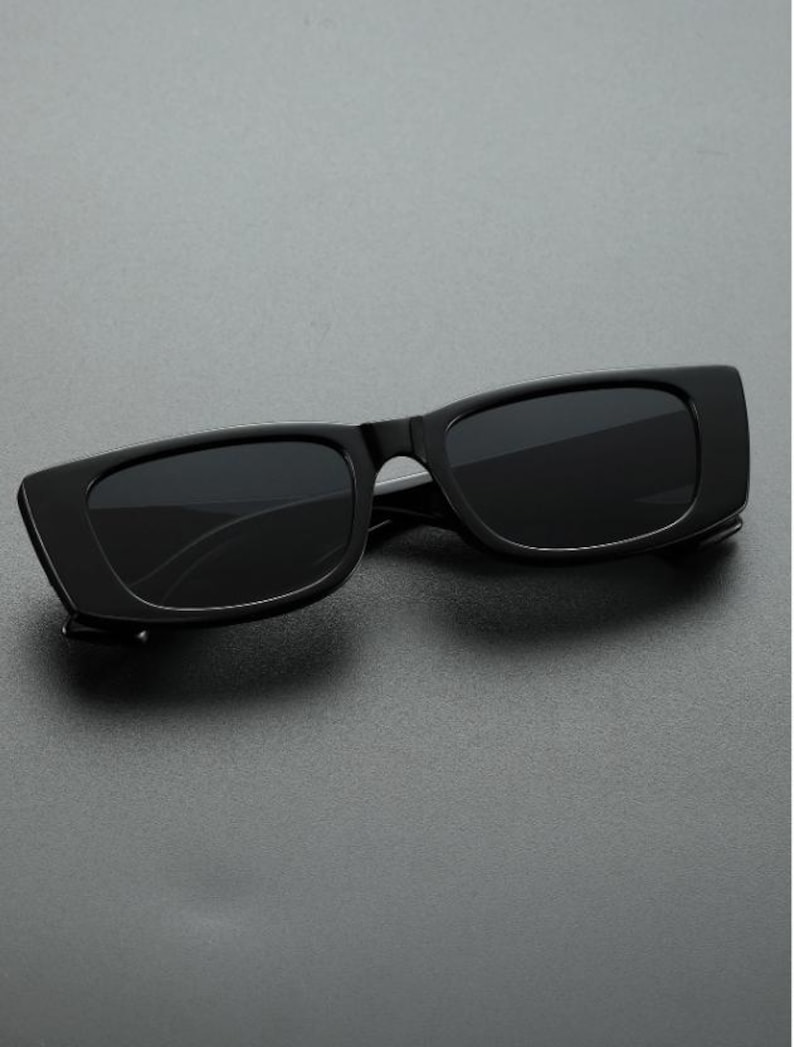 May include: Black rectangular sunglasses with dark lenses. The sunglasses have a thick, solid black frame and a sleek, modern design. The lenses are dark, providing shade and protection from the sun.