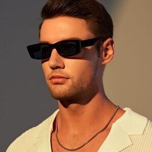 May include: Black rectangular sunglasses with dark lenses. The sunglasses are worn with a white collared shirt and a silver chain necklace. The image is lit with warm, natural light.