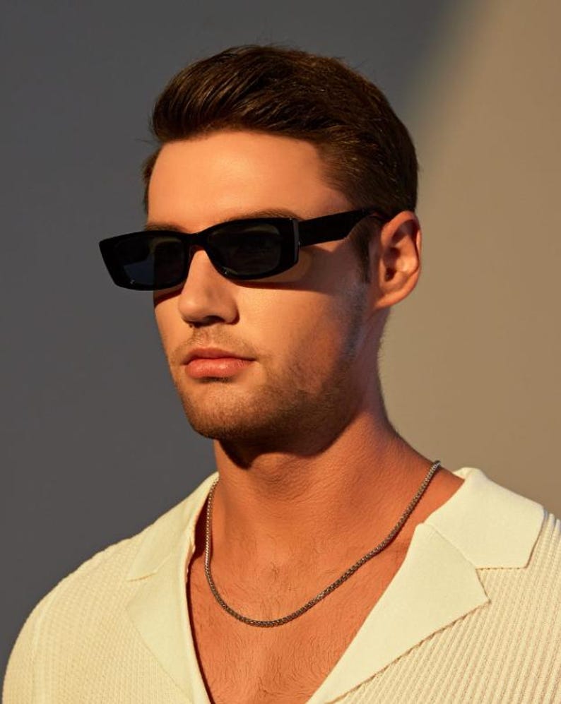 May include: Black rectangular sunglasses with dark lenses. The sunglasses are worn with a white collared shirt and a silver chain necklace. The image is lit with warm, natural light.