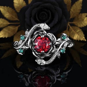 May include: A silver ring with a central red gemstone, encircled by a floral design and embellished with small green and white stones. The ring incorporates a snake motif winding around the floral setting. The backdrop features a black rose and gold leaves.