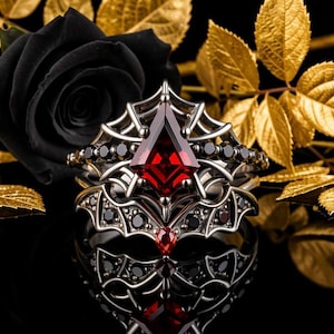 May include: A silver-toned ring featuring a central kite-shaped red gemstone, surrounded by a spiderweb-inspired metal framework. The band is accented with small, round black stones and a single small red teardrop-shaped stone at the base.