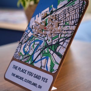 May include: A 3D relief map of Cleveland, Ohio, featuring a small red heart marker. The map is mounted on a wooden stand with a white plaque that reads: THE PLACE YOU SAID YES, THE ARCADE, CLEVELAND, OH. The map displays blue water, green areas, and white buildings.