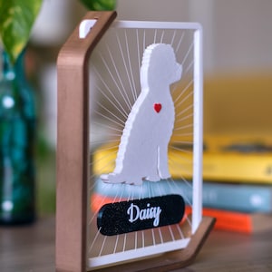 May include: A wooden framed dog silhouette art piece. The white dog silhouette has a red heart and is set against a white background with radiating lines. The dog's name, "Daisy", is on a black oval at the bottom.