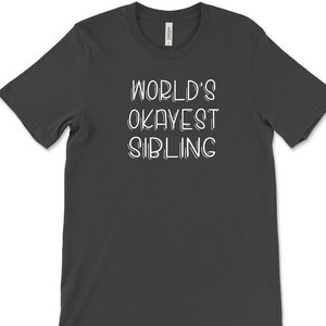 May include: Dark gray t-shirt with the white text "WORLD'S OKAYEST SIBLING" in a playful font. The shirt is a crew neck style and appears to be made of a soft material.
