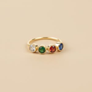 May include: A gold ring featuring four bezel-set gemstones in white, green, red, and blue. The ring has a simple, elegant design, with the gemstones evenly spaced along the band. The ring is set against a neutral background.