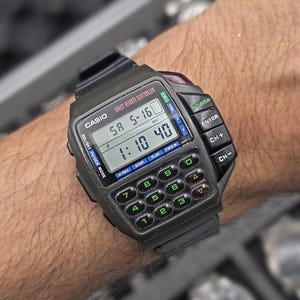 Op de afbeelding: Een zwarte Casio Wrist Remote Controller-horloge met een rekenmachinetoetsenbord en een digitale display met datum en tijd. Het horloge heeft knoppen voor stroom, kanaalbediening en volume. Het horloge zit op een zwarte polsband.