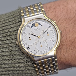 May include: A Seiko Quartz wristwatch with a silver and gold-tone bracelet and case. The watch face is white with Roman numerals and three sub-dials. The watch displays the day, date, and moon phase.