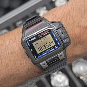 Op de afbeelding: Een zwarte Casio digitale horloge met een rechthoekige wijzerplaat en meerdere knoppen. Het horloge geeft de datum en tijd weer. De tekst "WAIST REMOTE CONTROLLER" is zichtbaar op de wijzerplaat.