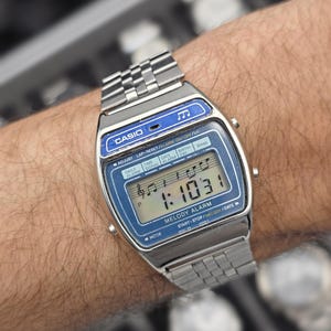 May include: A silver Casio digital watch with a blue face and a metal strap. The watch displays the time as 1:10:31 and features the text "MELODY ALARM". The watch is worn on a wrist.