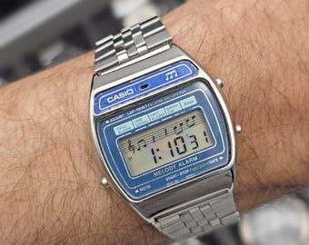 Puede incluir: Un reloj digital Casio plateado con esfera azul y correa metálica. El reloj muestra la hora 1:10:31 y tiene el texto "MELODY ALARM". El reloj está en una muñeca.
