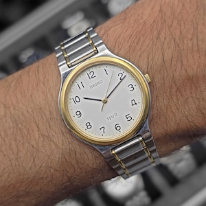 May include: A silver and gold Seiko Spirit wristwatch with a white face and black hands. The watch has a gold bezel and a silver and gold link band. The watch face displays the word "SEIKO" and "spirit".