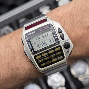 Op de afbeelding: Zilveren Casio Wrist Remote Controller horloge met een rekenmachine toetsenbord en digitale display. Het horloge geeft de tijd en datum weer. Het horloge heeft een rode accent op de bovenkant en zit om een pols.