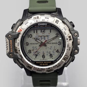 May include: A Casio digital watch with a black case and a green band. The watch face is gray with black numerals and hands. The watch has multiple buttons and features, including a compass and light.