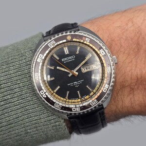 Puede incluir: Un reloj automático Seiko vintage con esfera negra, detalles dorados y visualización de día y fecha. El reloj tiene un bisel giratorio con números blancos y una correa de cuero negro. Es resistente al agua y tiene 18 rubíes.