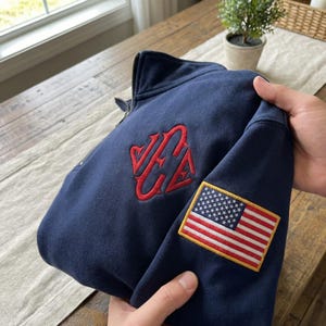 May include: A navy blue sweatshirt with a zip-up front, featuring a red embroidered design and an American flag patch. The sweatshirt has a high collar and a zipper. The chest design appears to be initials.