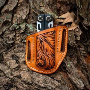May include: A tan leather sheath featuring intricate floral tooling and white stitching, designed to hold a black multi-tool. The leather has a smooth finish with dark brown accents in the recessed areas of the embossed leaf pattern.