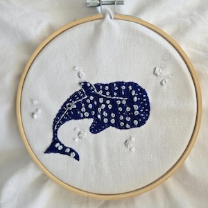 May include: Hand-stitched embroidery art featuring a blue whale with white spots, set within a wooden embroidery hoop. The whale is centered on a white fabric background, with small white details around the whale, suggesting bubbles.