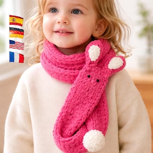 Puede incluir: Un niño lleva una bufanda rosa brillante con diseño de conejo. La bufanda tiene detalles blancos en las orejas y un pompón blanco. El niño lleva un suéter color crema. Accesorio infantil.