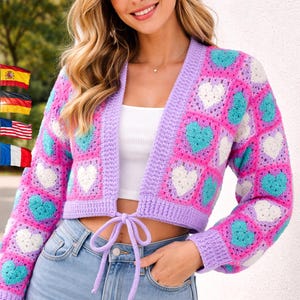 May include: Crochet cropped cardigan in shades of pink, purple, white, and teal. The open-front sweater features a heart pattern and a tie closure at the waist. The sleeves are long and the cardigan is worn with blue jeans.
