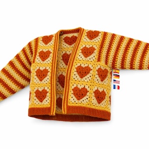 Może przedstawiać: Ręcznie robiony kardigan w stylu szydełkowym. Wzór przedstawia serca pośrodku i paski na rękawach. Kolorystyka obejmuje odcienie pomarańczowego, żółtego i brązowego. Kardigan prezentowany jest na białym tle. Wymiary w centymetrach.