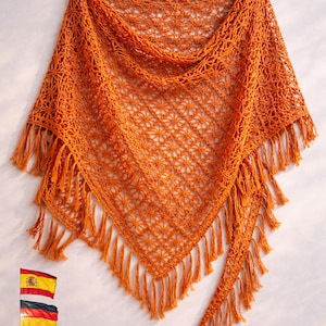 Puede incluir: Un chal naranja quemado, de forma triangular, con flecos en los bordes. El chal está hecho de un material suave y texturizado con un delicado patrón calado. El chal se muestra sobre un fondo claro.