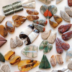 May include: A selection of polished gemstones in diverse shapes and colours, including teardrops, triangles, and ovals. The stones display a range of patterns, from striped to speckled, with colours like red, green, brown, and white. The gemstones are arranged on a white surface.