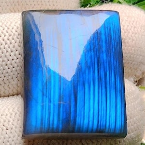 Può includere: Una gemma rettangolare lucida, di colore blu vibrante con striature verticali. La pietra è tenuta in un tessuto beige, con una moneta sullo sfondo, su uno sfondo di erba verde.